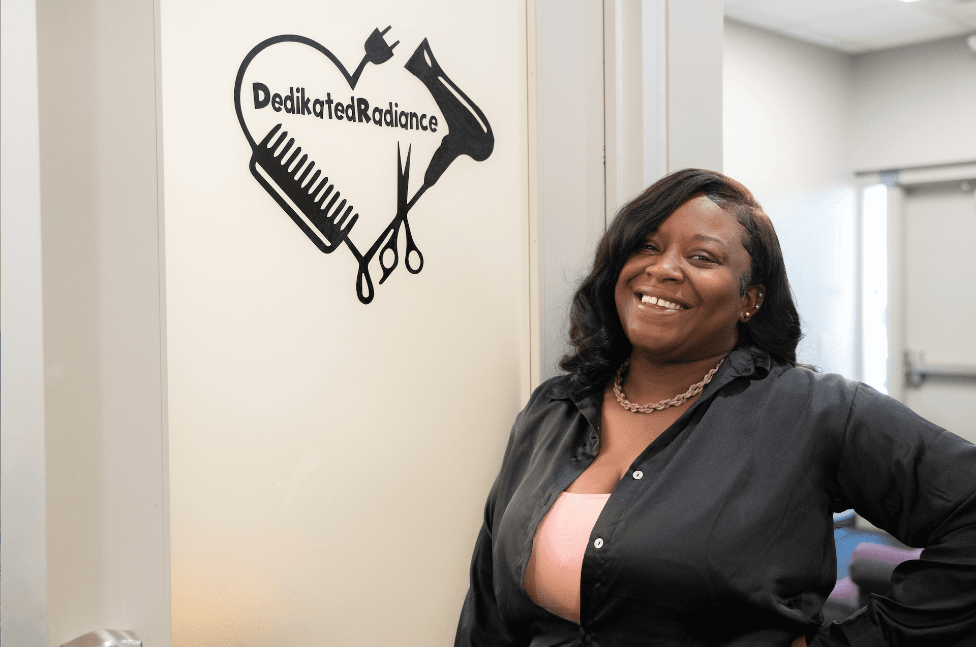 Independent hair stylist smiling beside her branded suite door in a Florence salon suite rental at The Hive Salon Studios, building her client base.